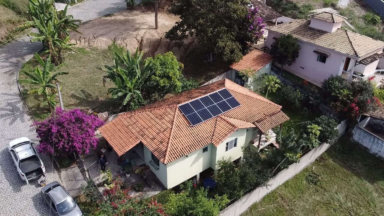 Vista aérea de casa verde com telhado laranja e painéis solares, cercada de vegetação e flores